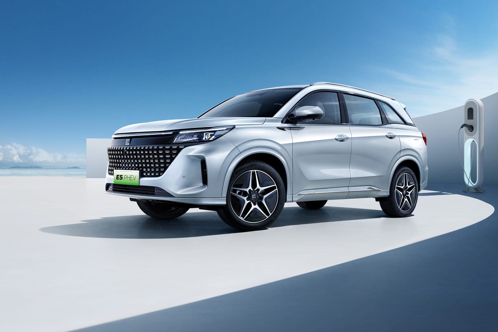 La DFSK E5 PHEV argento, di tre quarti, accanto a una colonnina di ricarica